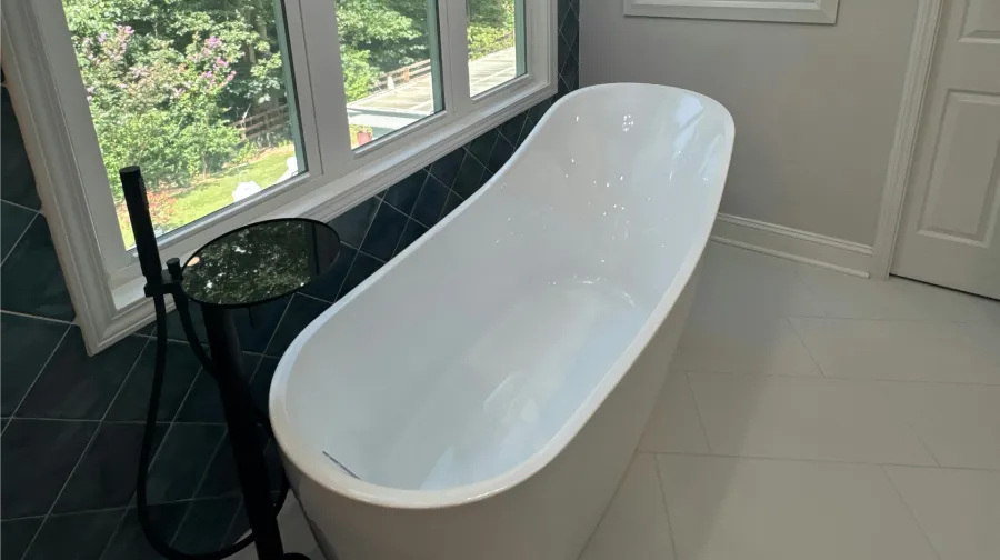 A modern bathroom features a white freestanding bathtub, large window overlooking trees, and a crystal chandelier.